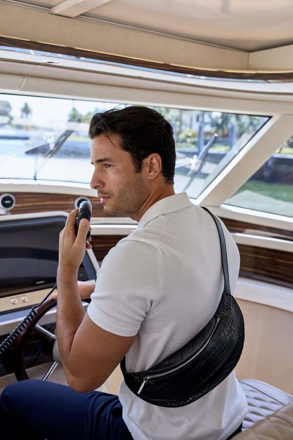 Man in white polo at boat's helm holds a radio and wears a black crossbody bag during a Miami photoshoot