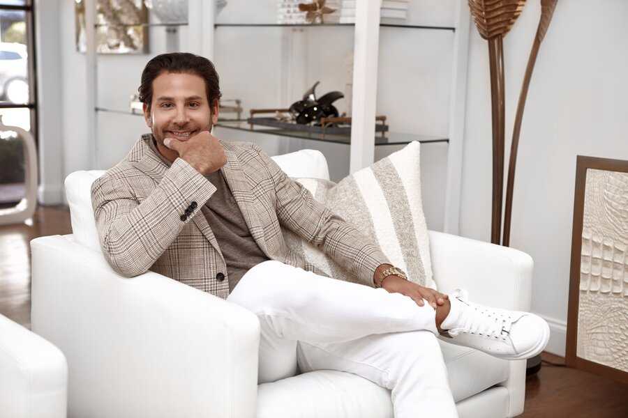 Man in a plaid blazer and white pants sits smiling on a white armchair during stylish Miami photoshoot