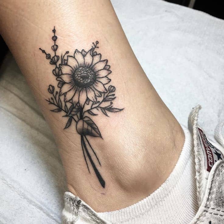medium-sized black and grey tattoo on woman's ankle of a bouquet of a sunflower with smaller flowers and stems