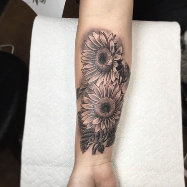large black and grey tattoo on man's forearm of a two realistic sunflowers with leaves around them