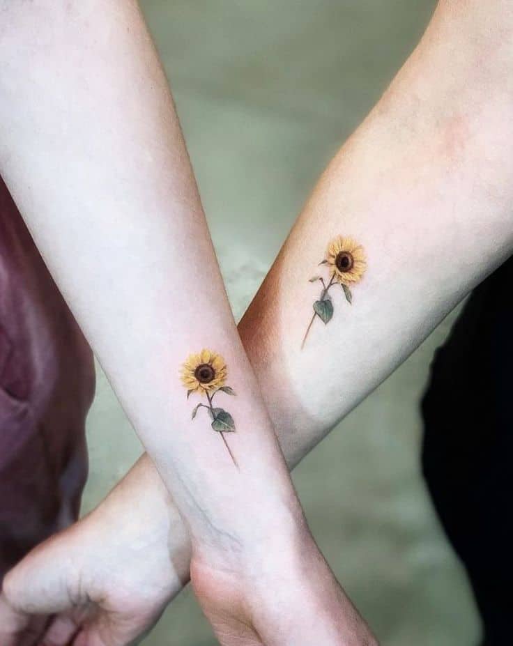 small matching color tattoos on two women's forearms of realistic sunflowers with stems