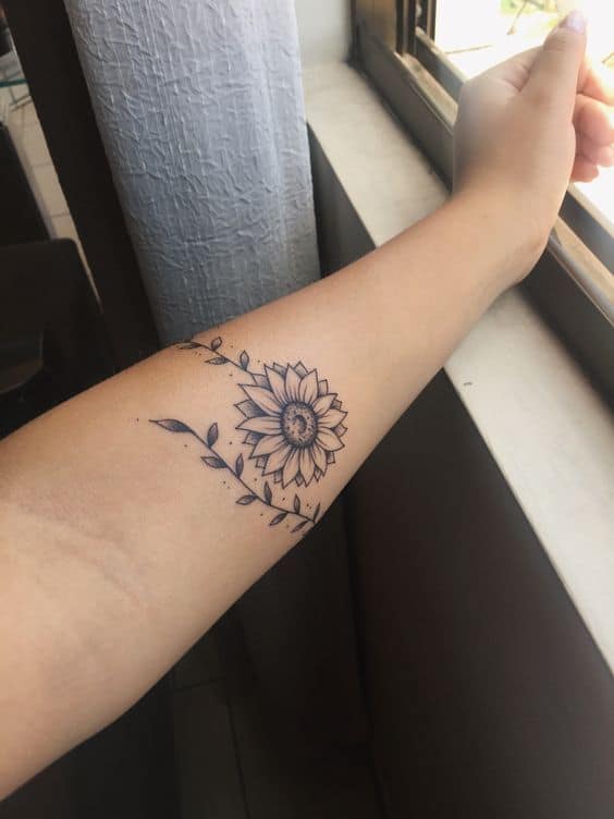 medium-sized black and grey tattoo on woman's forearm of traditional sunflower with vine wrapping around arm