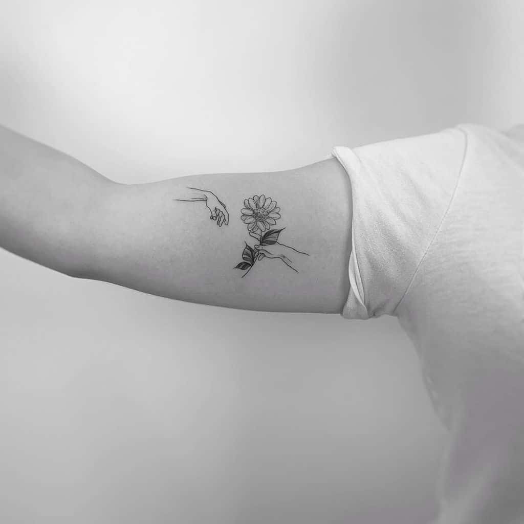 small black and grey tattoo on woman's upper arm of michaelangelo's creation of adam with a sunflower in hands