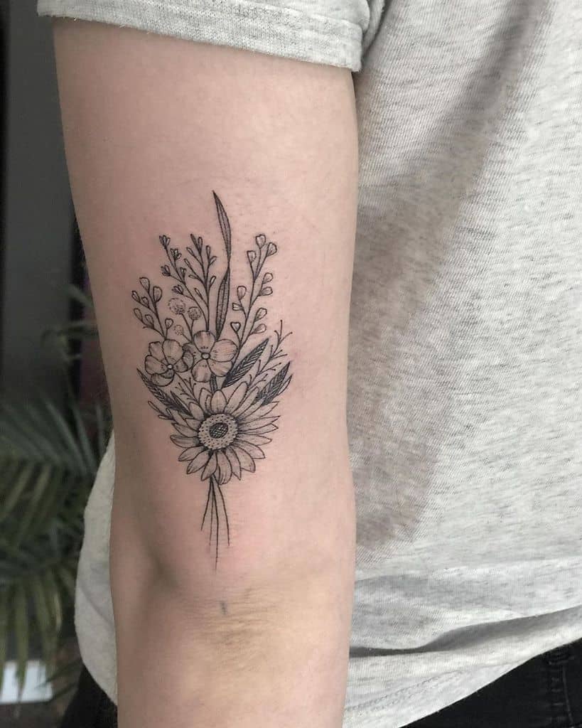 medium-sized black and grey tattoo of a sunflower and pansey bouquet on back of woman's upper arm