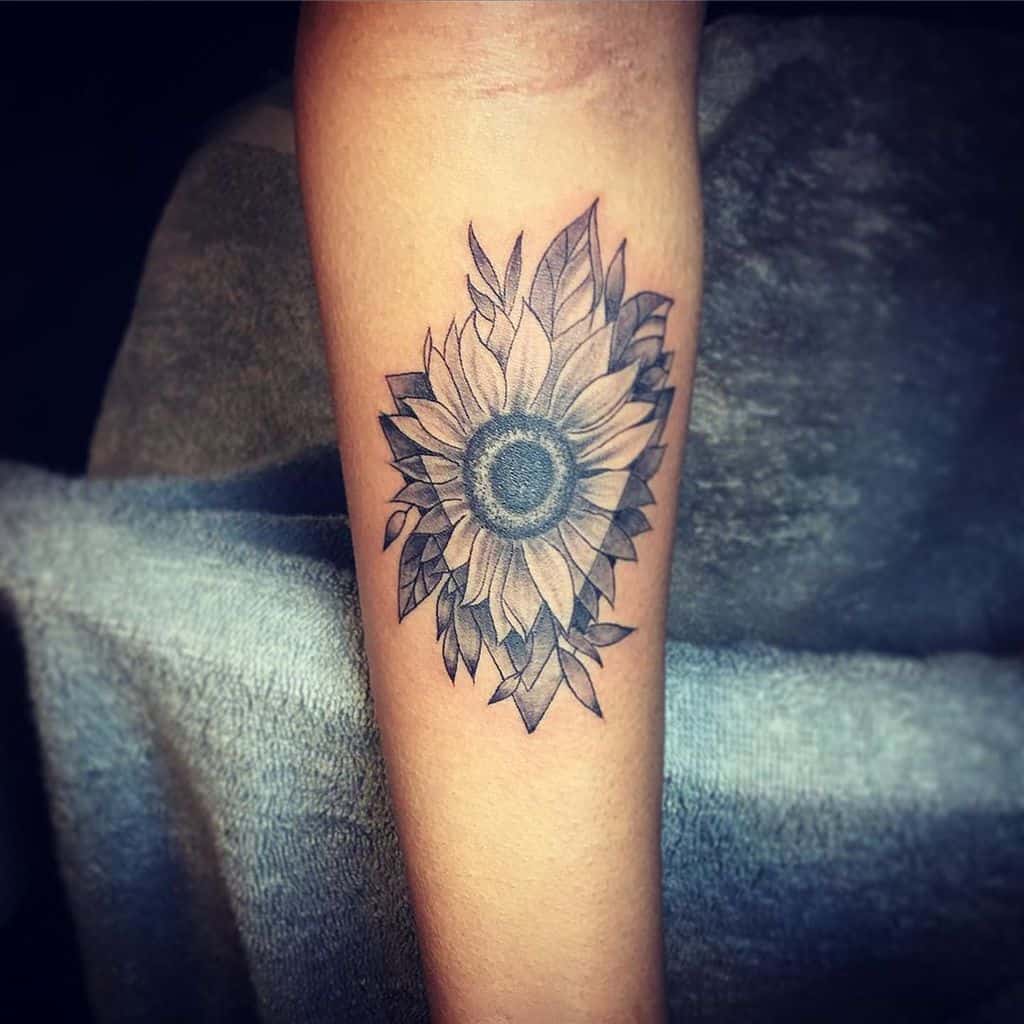 large black and grey tattoo on forearm of realistic sunflower inside geometric triangle and leaves around it