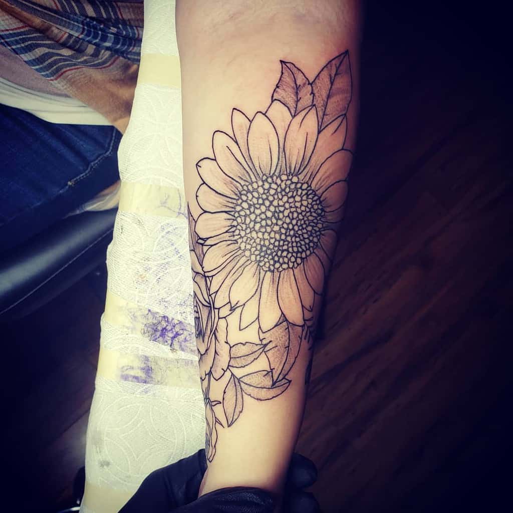 large black and grey surrealism tattoo on woman's upper arm of a sunflower with stem