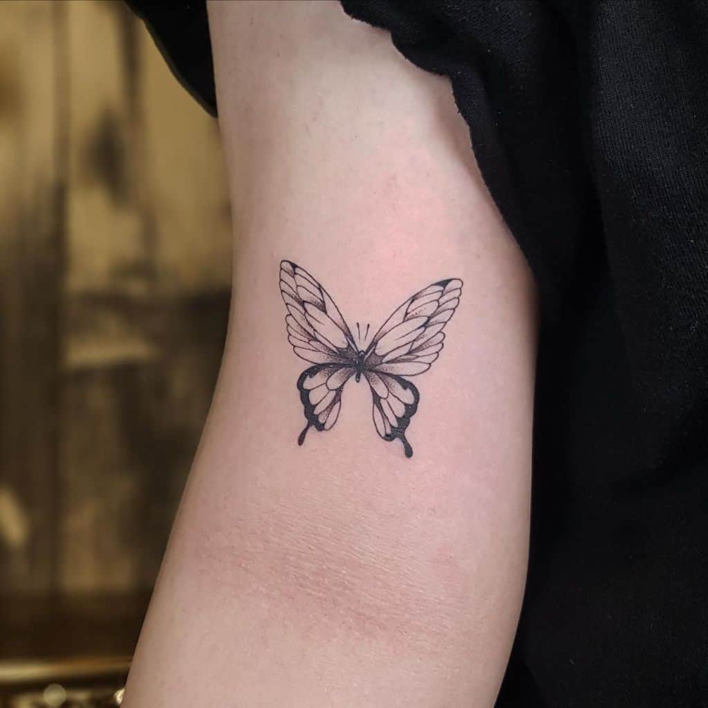 small black and grey tattoo on woman's upper arm of realistic delicate butterfly