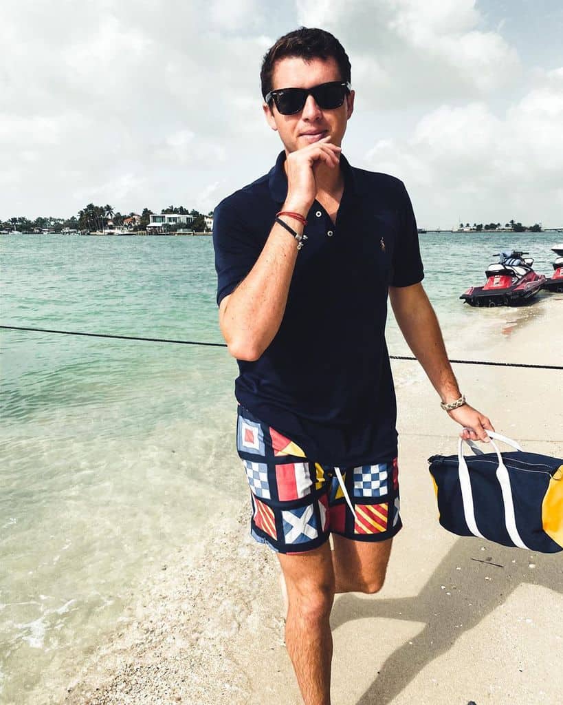 A Man On The Beach In Patterned Swim Trunks And A Classic Navy Blue Polo