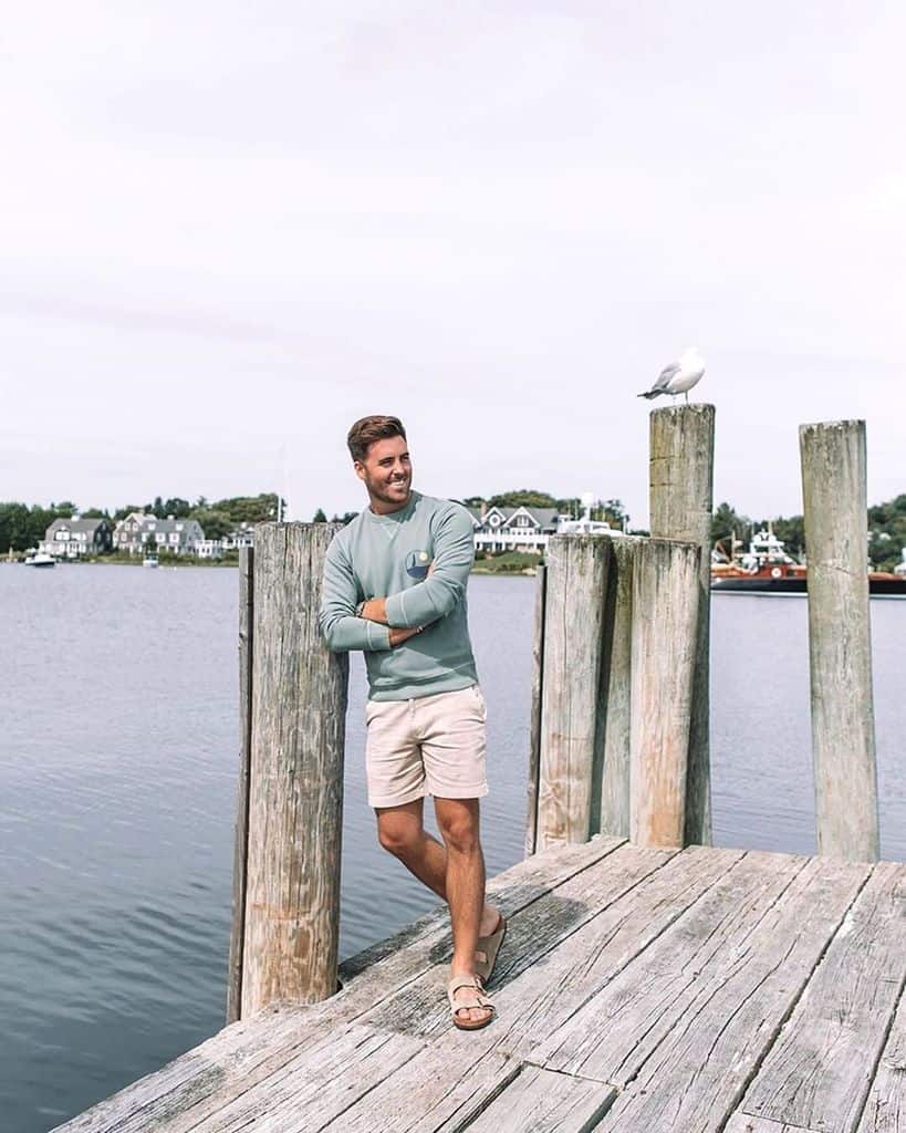 A Man Wearing A Bluish Green Crewneck Sweater While Standing On A Pier With His Arms Crossed