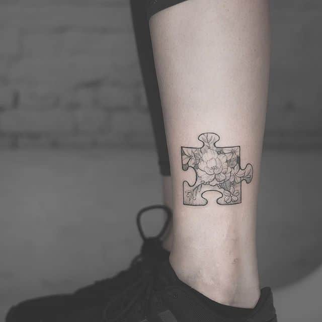 Black and gray ankle tattoo of puzzle piece with realistic flowers inside.