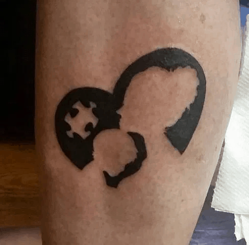 Black tattoo of a heart with negative puzzle piece and mother and child silhouette.