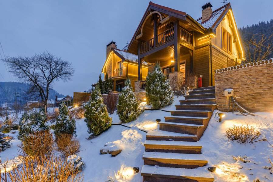 Wooden house with warm lights and curb appeal, nestled among snow and trees, steps rising from a snowy garden at dusk