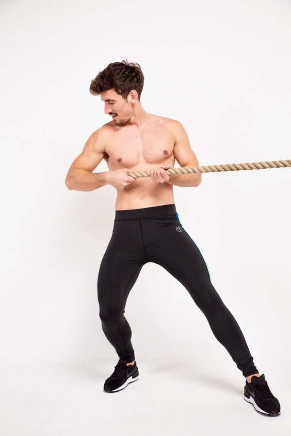 Shirtless man wearing compression pants and sneakers stands wide, pulling a thick rope against a white background