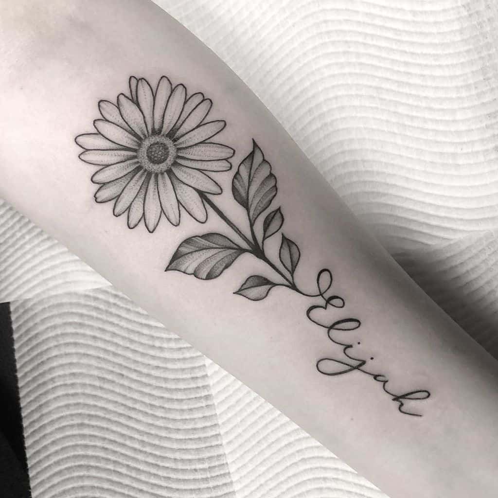 Forearm tattoo traditional black and grey stipple shading daisy with stem forming cursive script ‘Elijah
