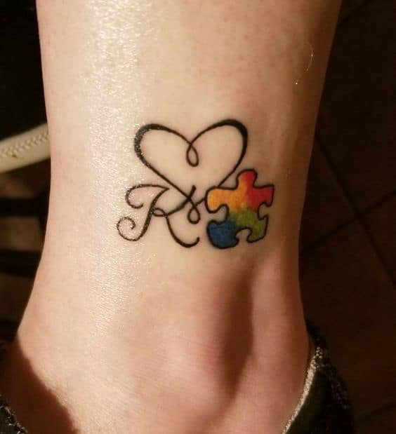 Full color ankle tattoo of a cursive “K”, heart and multi-colored puzzle piece. 