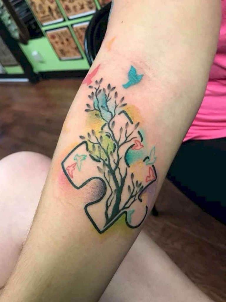 Full color forearm tattoo of a puzzle piece with a multi-colored tree and flying birds.