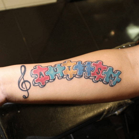 Full color forearm tattoo of a treble clef and puzzle pieces spelling “AUTISM”.