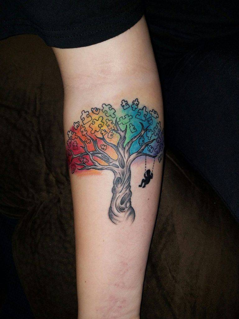 Full color forearm tattoo of a tree with multi-colored puzzle piece leaves and a child on a swing.