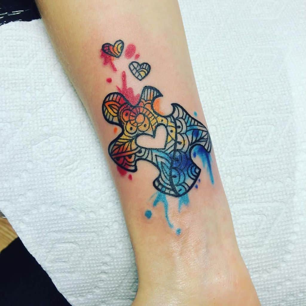 Full color forearm tattoo of puzzle piece with tribal designs, watercolor splashes and negative space heart.