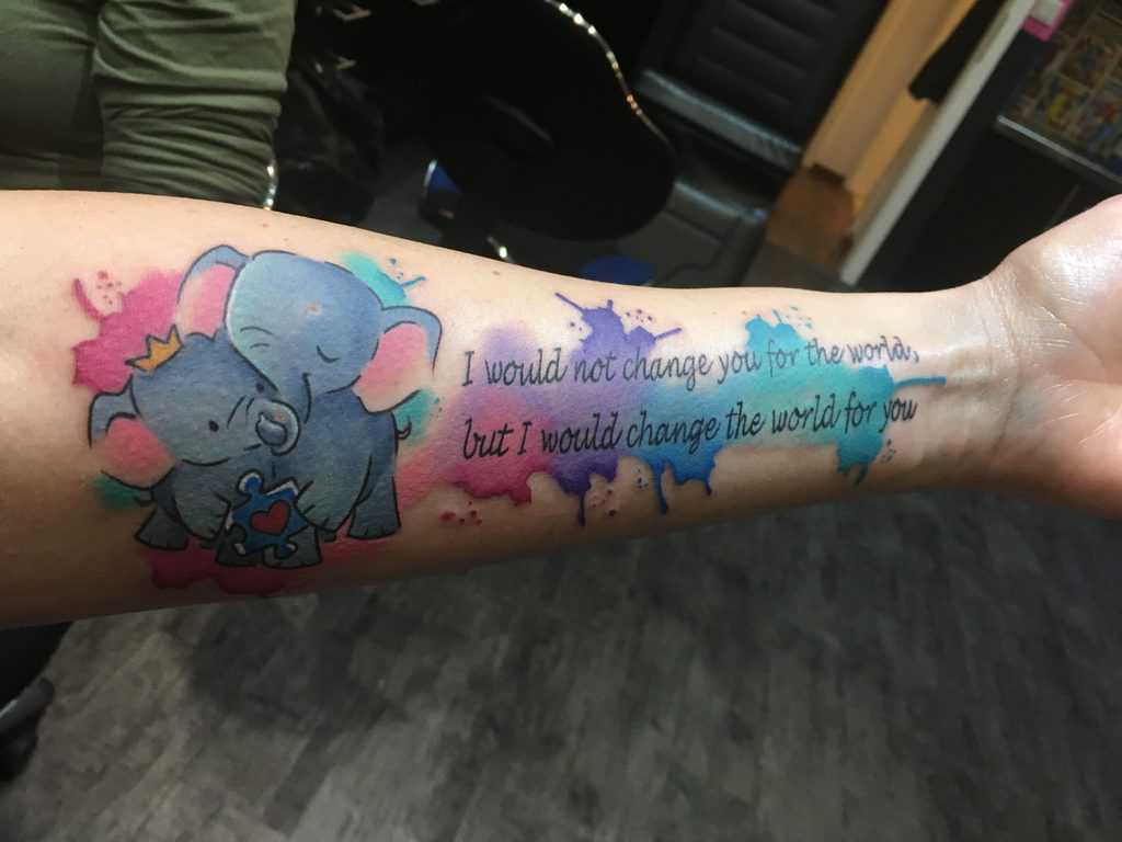 Full color forearm tattoo of two elephants holding a puzzle piece and watercolor splashes with text.
