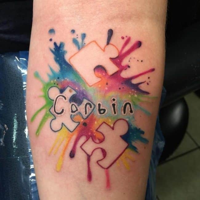 Full color forearm tattoo of watercolor splashes, negative space puzzle pieces and child-like “Corbin” script. 