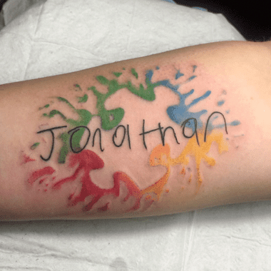 Full color forearm tattoo of watercolor splashes a negative space puzzle piece and child-like script: “Jonathan”.