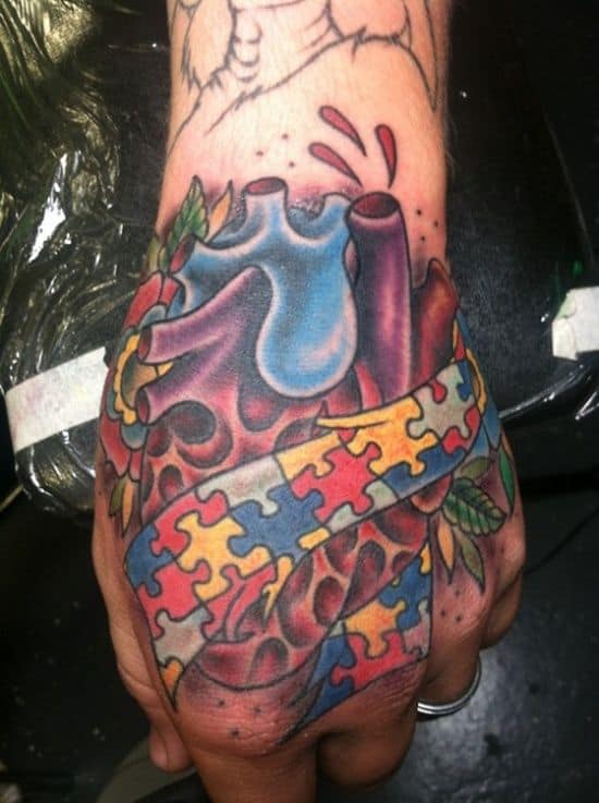 Full color hand tattoo of anatomical hear wrapped in multi-colored puzzle piece scrolls. 