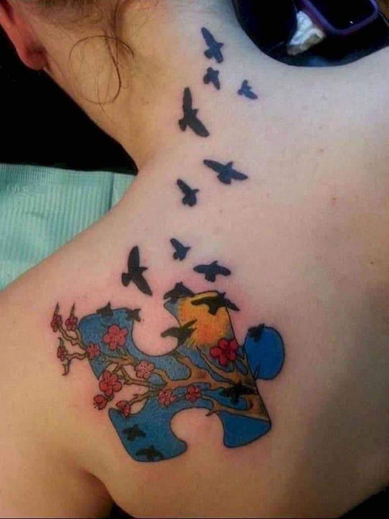 Full color shoulder blade tattoo of a puzzle piece with a sun, blue sky, cherry tree and birds flying.