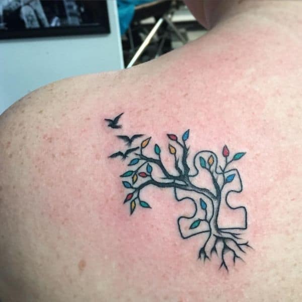 Full color shoulder blade tattoo of tree with multi-colored leaves growing over a puzzle piece and birds flying.