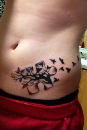 Limited color stomach tattoo of a puzzle piece with a multi-colored tree and black birds flying.