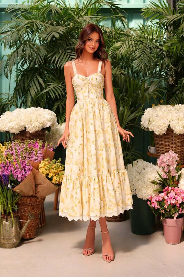 Woman in a yellow floral OhPretty sundress showcases timeless style, smiling among bouquets in a bright indoor garden setting