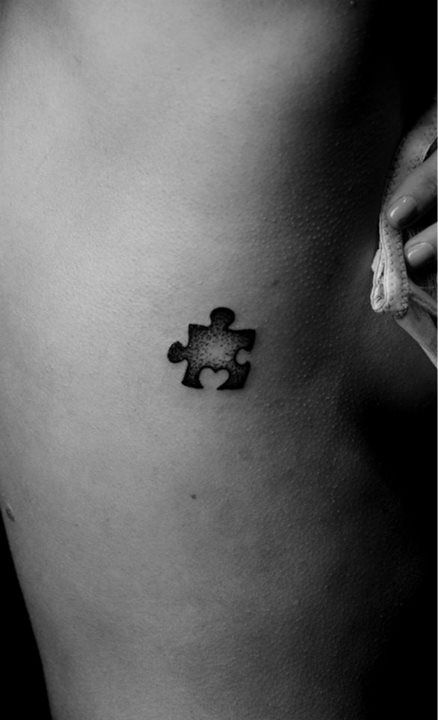 Small black and gray rib tattoo of a puzzle piece with stipple shading.