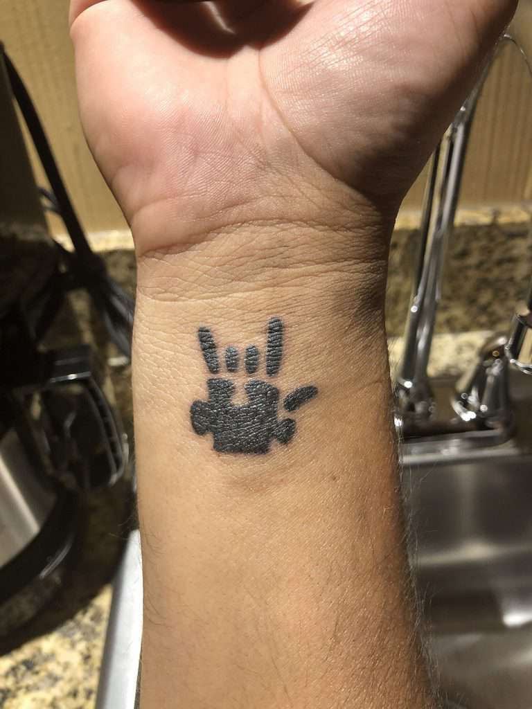 Small black and gray wrist tattoo of a puzzle piece made to look like the hand symbol for love.