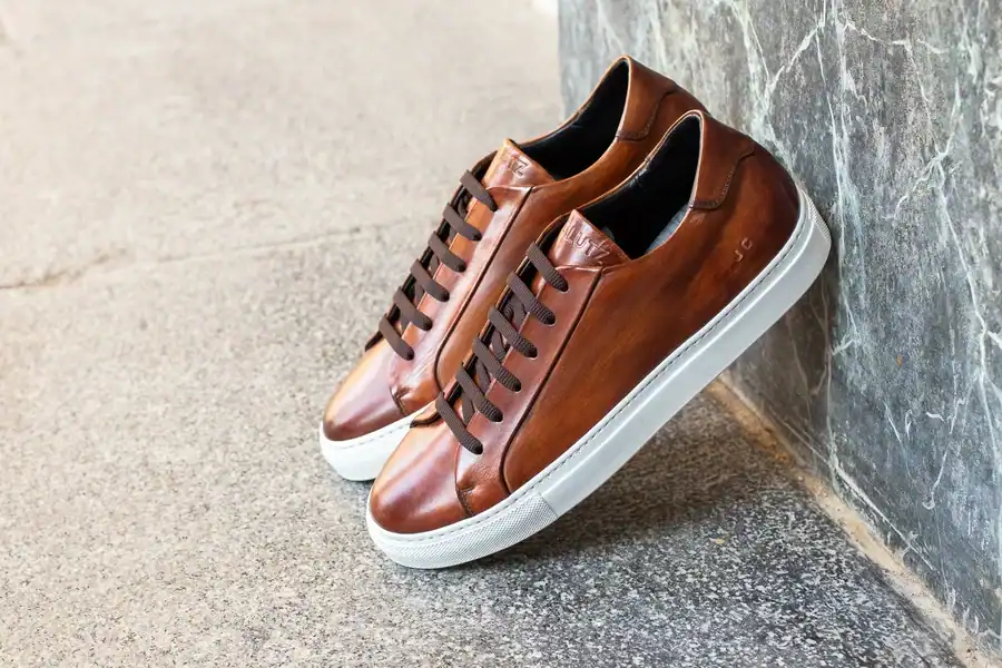 A pair of brown high-quality J.C.LUTZ leather shoes with white soles rest against a gray textured wall