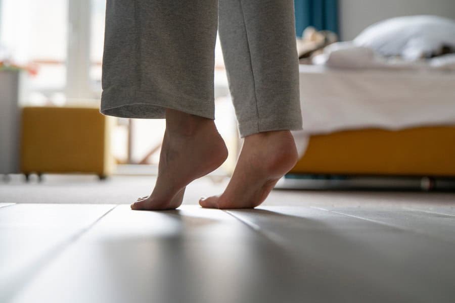 Person standing on tiptoes on a wooden floor, wearing gray sweatpants, with a blurred bed in the background