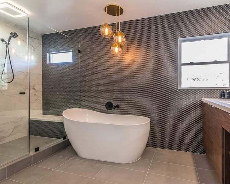 Gold globe pendants above a white freestanding tub with dark penny tile walls and a glass-enclosed marble shower in a modern bathroom.