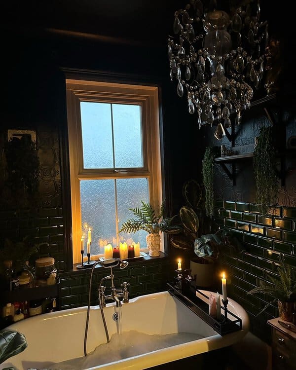 Crystal chandelier above a white clawfoot tub with dark green tile, candles, and lush plants in a moody bathroom setting.