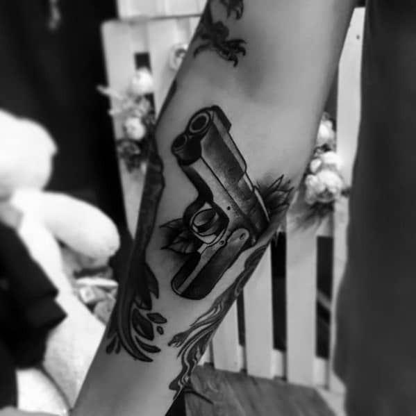 Realistic black and gray tattoo of a 1911 pistol on the forearm, featuring detailed shading and floral elements for a bold yet artistic look.