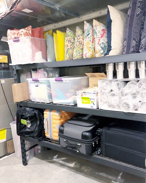 Open basement shelves with pillows, storage bins, luggage, and disposable cups neatly organized.