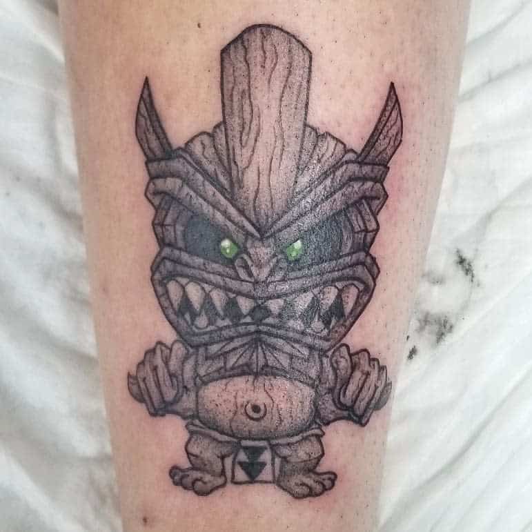 Black and grey Tiki tattoo featuring a cartoonish mask with sharp teeth, pointed ears, and green eyes with arrow down in his clothes