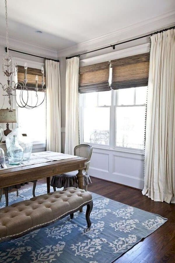 Farmhouse dining room with white curtains, woven shades, a rustic wooden table, and a tufted bench.