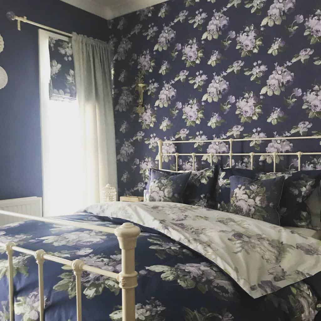 Bedroom with dark floral wallpaper, white metal bed frame, and matching floral bedding.