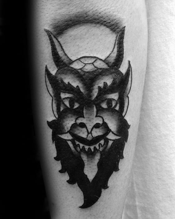 Blackwork Krampus tattoo featuring a grinning horned face with bold shading, sharp fangs, and a circular background accent.