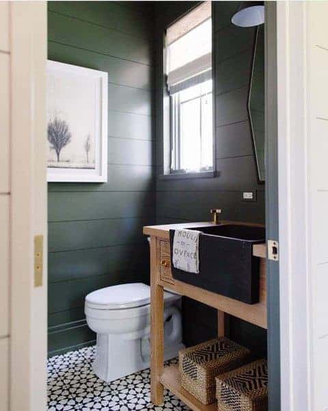 Rustic half-bath with dark green shiplap walls, wooden vanity, and patterned floor tiles.