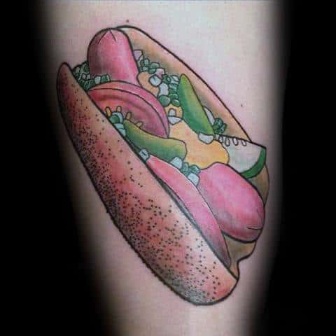 Hot dog tattoo featuring a sausage with mustard, onions, tomatoes, and pickles in a poppy seed bun on the forearm.