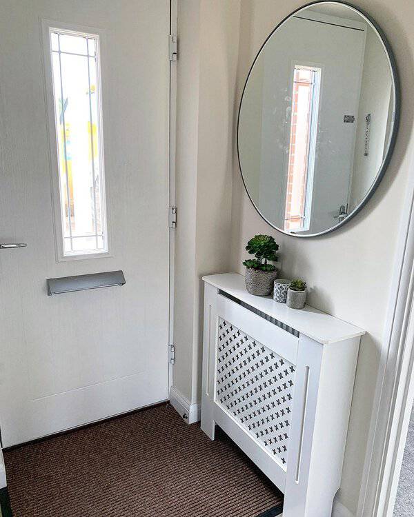 Round mirror in a bright entryway above a white radiator cover with potted plants, reflecting natural light for an open and airy feel.
