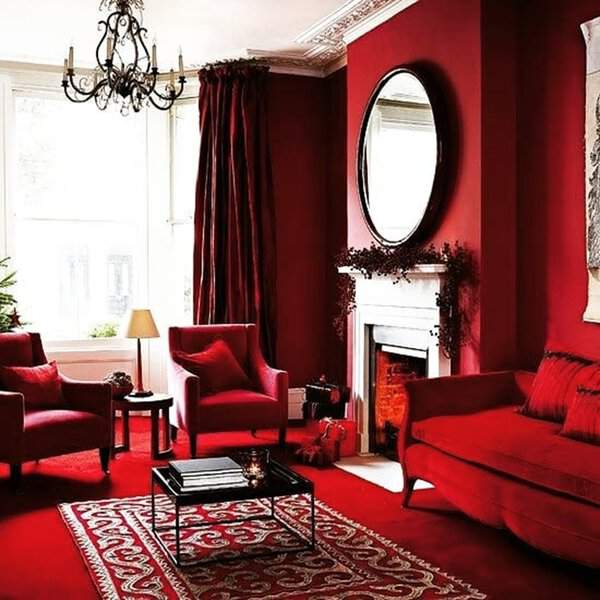 Round mirror above a white fireplace in a bold red living room with velvet furniture, chandelier, and patterned rug for a dramatic look.
