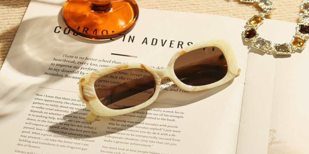 Beige prescription sunglasses, an orange glass, and a jeweled necklace lying on an open book under warm sunlight