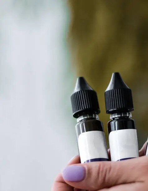 A hand with purple nail polish holds two small bottles for hobbyist vaping, each featuring black caps and blank white labels