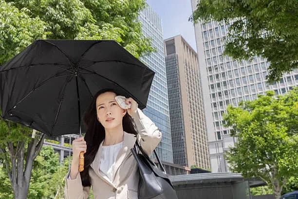 Woman in a suit wipes her face with a handkerchief, shielding her healthy skin beneath a black umbrella on a sunny city day
